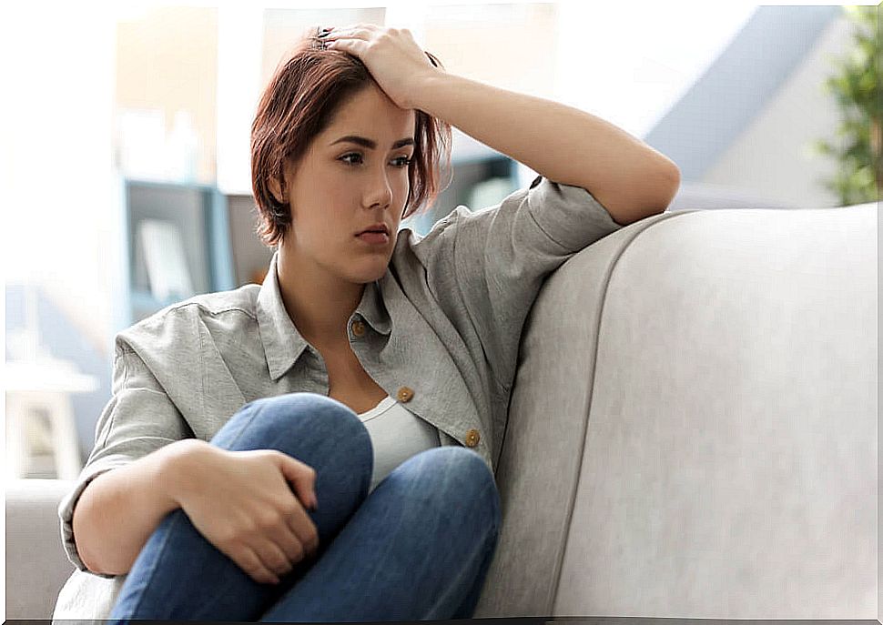 Young woman surrounds her legs with one hand and rests the other on her head thoughtfully while sitting on the sofa.